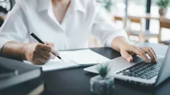 Persoon werkt op laptop en schrijft aantekeningen op papier, moderne kantooromgeving.